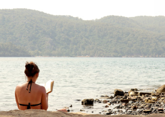 Fotografia de mulher de bikini, na praia, de costas para a camera, a ler um livro.