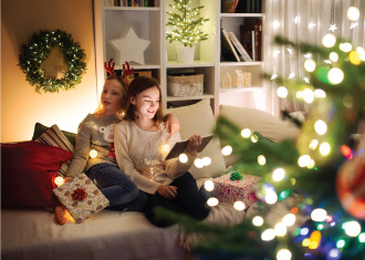 Mãe e filha sentadas no sofá, a ler um livro, com árvore de natal no fundo.