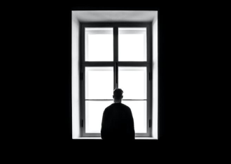 Homem vestido de preto, a olhar para a janela com luz solar, no meio da escuridão.